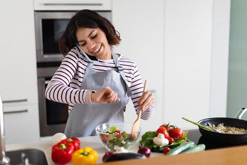 Cheerful young brunette pretty middle eastern woman wearing apron having phone conversation, looking at watch on her wrist, checking time while cooking dinner at home, waiting for guests, copy space