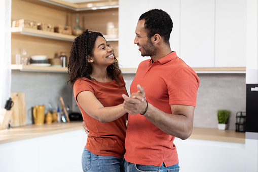 Smiling handsome young black husband and wife in red t-shirts dancing, enjoy free time and music, have fun together at kitchen interior. Anniversary, date, love, romance and relationships at home