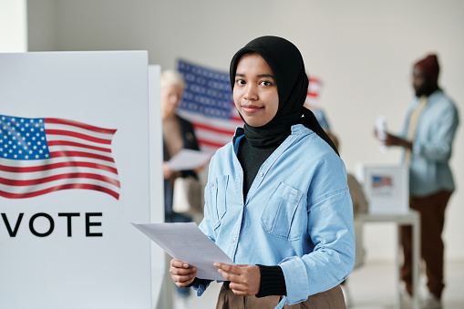 Young smiling Muslim woman in hijab holding ballot paper and looking at camera while standing against voters in polling place