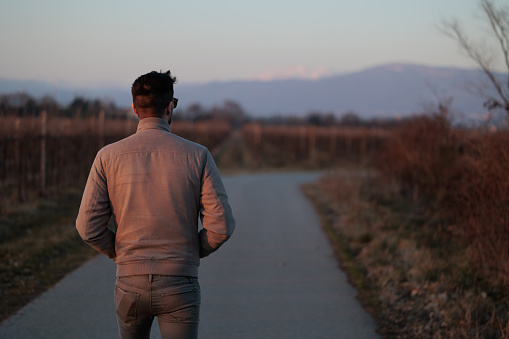 portrait of a young adult man, seen from the back, from the head to the waist, walking along a road in a natural countryside environment in Slovenia, at sunset in winter