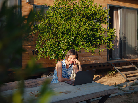 Happy young asian woman with laptop resting outdoors in tiny house in summer garden, weekend away and remote office idea, early morning. Tiny houses and Small Living concept.