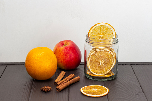 Fresh apple and orange, cinnamon sticks and dried orange slices in a glass jar. Copy space. Dark wooden and white background.
