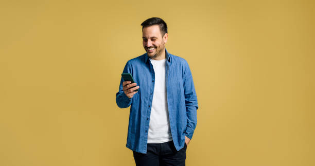 istock Smiling mid adult handsome man browsing social media on smart phone while posing with hand in pocket on isolated yellow background 1473027254