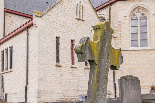 Old stone catholic cross with the exterior of a catholic church in the background.