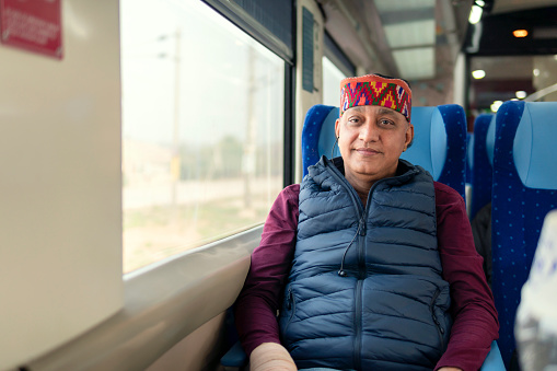 Portrait of Happy senior man of Himachal Pradesh in typical Himachali cap traveling by train. He is sitting near the window and looking and the camera with a smile.
