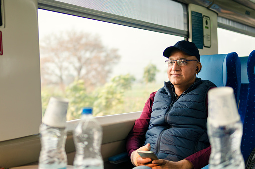 Senior Indian man traveling in train and listening music on smartphone with earphones and enjoying the view.