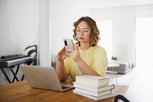 Young woman sitting at table using mobile phone while working on laptop at home. Caucasian female texting on her phone while working from home.