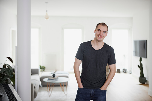 Portrait of a handsome young man standing with hands in pocket at home. Confident male standing casually in living room and looking at camera.