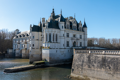 Horizontal landscape photo of residential buildings, a distant bridge and a blue sky and trees reflected in the calm water of a canal in the historic town of Azay-Le-Rideau, Loire Valley, France, in Spring.