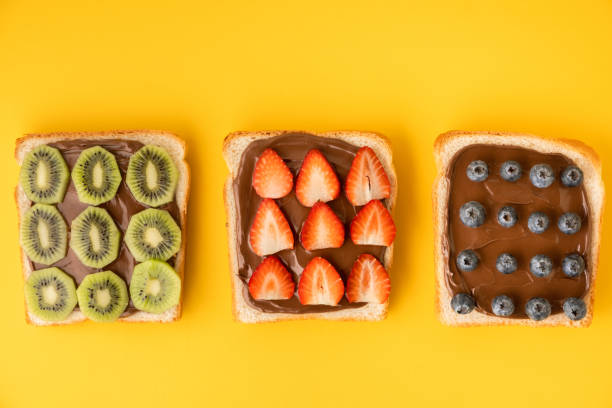 istock Top view of white toast bread slices with chocolate hazelnut spread decorated with fresh summer berries: strawberries, blueberries and kiwi fruit on the yellow background. Morning breakfast concept. 1473038921