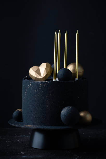istock Luxury anniversary cake with black velvet sprayed coating decorated with golden chocolate spheres, heart and candles on top. Vintage cake for beloved one on the black background 1473038927