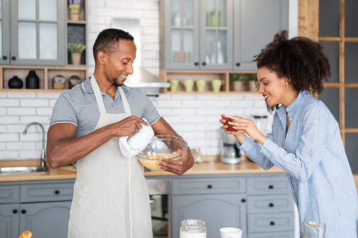 Couple cooking together, having fun time in the kitchen. Multiracial man mixing eggs in a bowl, while his girlfriend taking picture of him with her mobile phone.