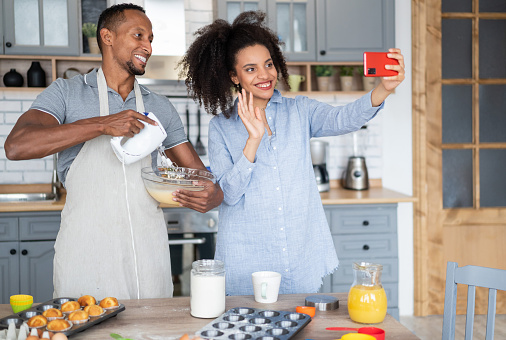 Couple cooking together, having fun time in the kitchen. Multiracial man mixing eggs in a bowl, while his girlfriend shooting with her mobile phone, taking a selfie, vlogging and waving