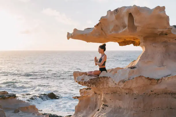 Young woman meditating on a cliff by the ocean at sunset