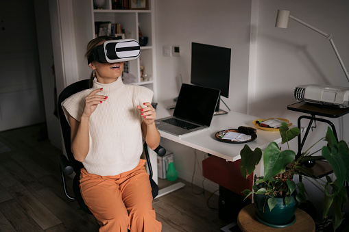Caucasian female gesturing while using virtual reality glasses. Woman wearing vr goggles sitting at home office desk.