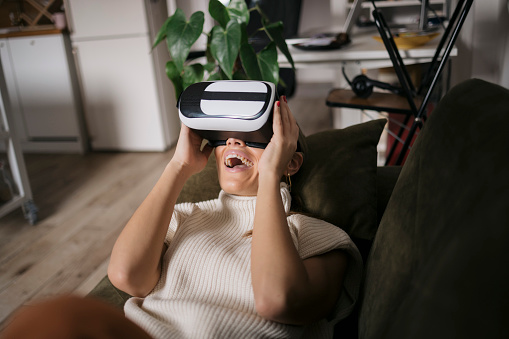Excited woman enjoying using vr glasses at home. Female lying on sofa wearing virtual reality simulator and laughing.