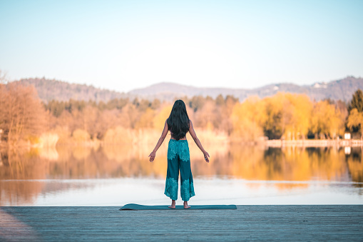Female New Ager with black, long hair over her back, standing at a lakefront with arms slightly away from the body, practicing breathing and meditation. Full length image, back view, blurred background.