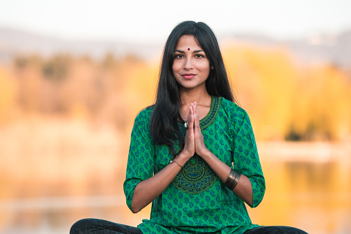 A portrait of a young, attractive Indian woman, sitting down in front of a large body of water, wearing an emerald green shirt and a bindi on her forehead, looking at the camera. Waist up image, blurred scenic background.