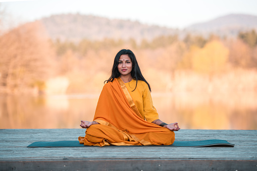 Young Indian female yoga teacher wearing an orange sari and a dot on the forehead, sitting comfortably while meditating and slightly smiling. Looking at the camera. Full length image, blurred bright background.
