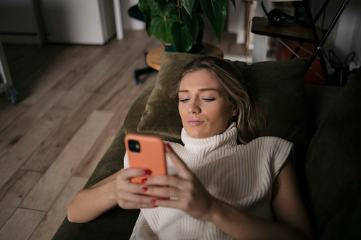 Beautiful young woman lying on living room sofa using mobile phone. Female relaxing at home on a weekend texting on cell phone.
