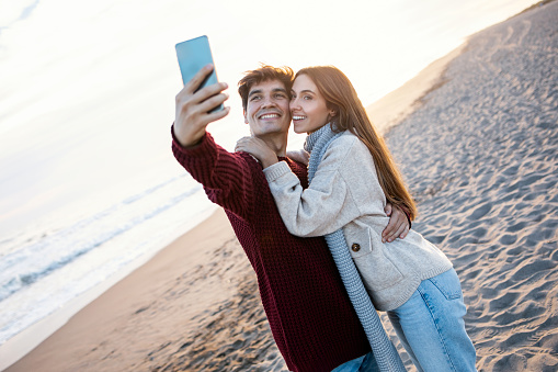Shot of beautiful young couple making photos with mobile phone in a cold winter on the beach.