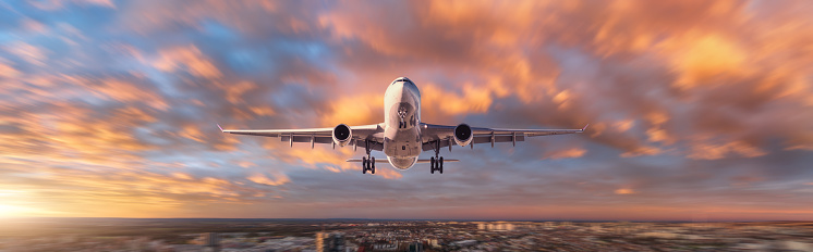 Plane is flying in colorful sky over the city at sunset. Landscape with passenger airplane, and blurred background with sky with orange clouds. Aircraft is landing. Front view. Travel. Aerial view