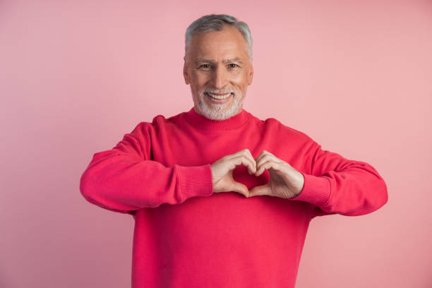 istock Older man shows a gesture from the heart. Man affectionate, making a heart with fingers on a pink background, place for text, copy space 1473055212