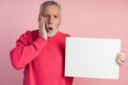 Surprised senior man holding a white sheet of paper. Isolated on pink background man holding blank white sheet of paper, copy space, place for advertising.