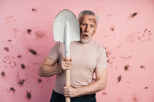 Surprised, the older man looks out from behind the wreck on a dirty, pink background. Elderly man isolated on copy space.