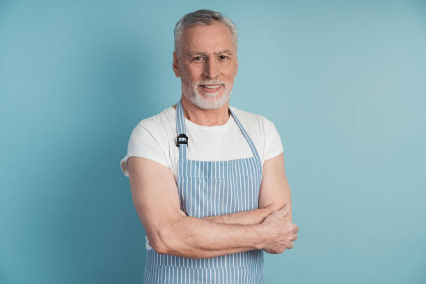 istock Attractive, elderly man in a blue apron posing on a blue background. Handsome man isolated on blank wall background 1473055509