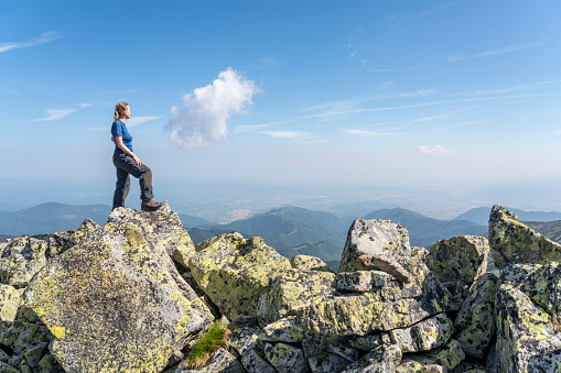 Mature woman standing on the rock against blue sk. The most beautiful trekking in Romania, the National park Retezat ( 2485 m) , Yellow lichen on the rocks in front view, in distance mountain range.