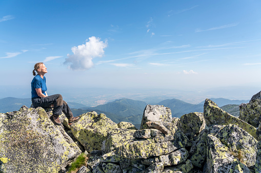 Mature woman sitting on the rock against blue sk. The most beautiful trekking in Romania, the National park Retezat ( 2485 m) , Yellow lichen on the rocks in front view, in distance mountain range.