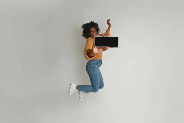 Black woman, casual dressed, on neutral background. Cheering and jumping in the air while holding a mobile in her hand.
Looking at camera