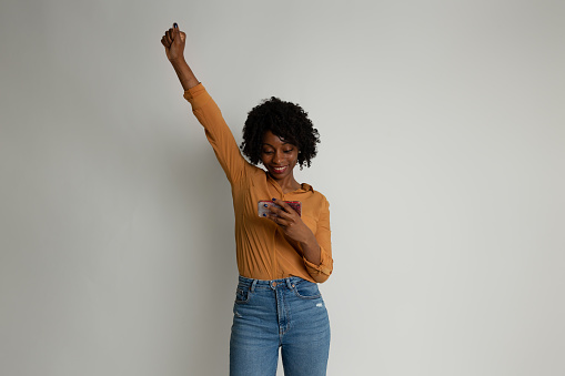 Black woman, casual dressed, on neutral background. Cheering while holding a mobile in her hand.
Looking at mobile