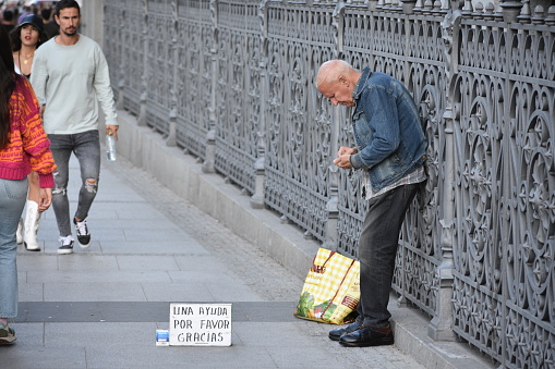 This photo captures an elderly homeless man sitting on a Madrid street, asking for help and money. He appears to be struggling and in need of assistance, as he displays a cardboard sign in the hopes of receiving some spare change from passersby. The image highlights the ongoing issue of homelessness and poverty that affects many people in cities across the world.