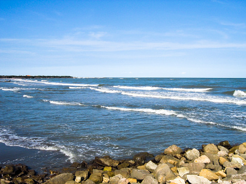 Ocean foamy waves roll onto the shore. The shore is strewn with stones. Bright day