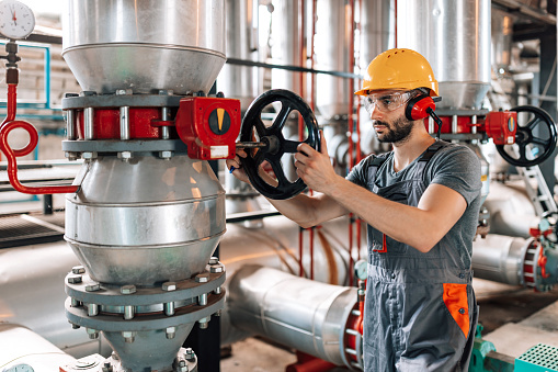 A young Caucasian male engineer in full safety equipment is turning the valve on one of the boilers in a heating plant.