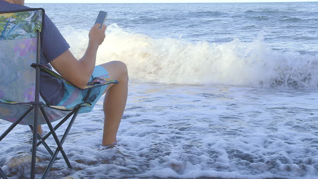Man resting in a portable chair on the seashore, waves touching his feet, man sitting with cell phone in hand