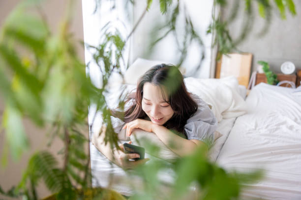 istock Young woman using smartphone on bed in the morning - lying on front, smiling 1473071710