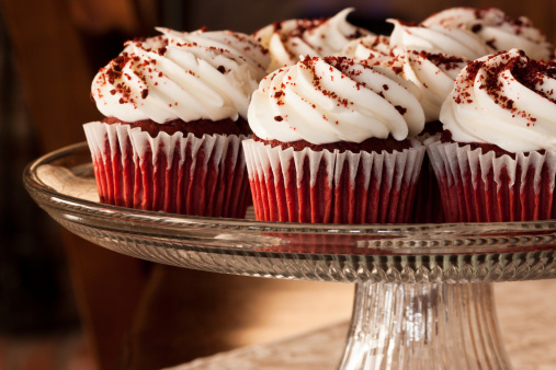 Stock photo showing close-up view of a batch of freshly baked, homemade Father Christmas hat design cupcakes, in paper cake cases, displayed on a red background. The cakes are topped with swirls of white and red butter icing. Home baking concept.