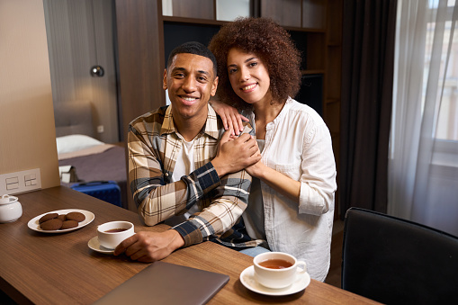 Beautiful multiracial young couple having morning coffee in hotel room, guys in casual clothes