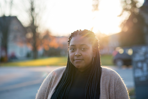 Shoulders up portrait of a beautiful black millennial woman with her hair in braids, wearing a turtle neck and beige cardigan looking at the camera while standing on the sidewalk of a street. She has a neutral, pleasant expression on her face.