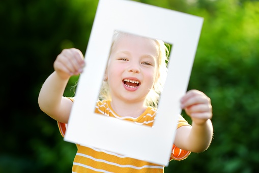 Cute cheerful little girl holding white picture frame in front of her face. Adorable child framing her smiling face.