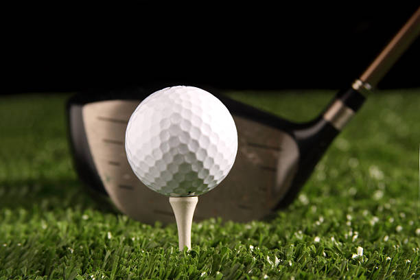 White golf ball on tee with a 1 wood driver Close up white golf ball on white tee secured in green grass(artificial turf) with 1 wood driver (golf club) behind the ball ready to be hit, black backgroung, space for copy to be added. night golf stock pictures, royalty-free photos & images