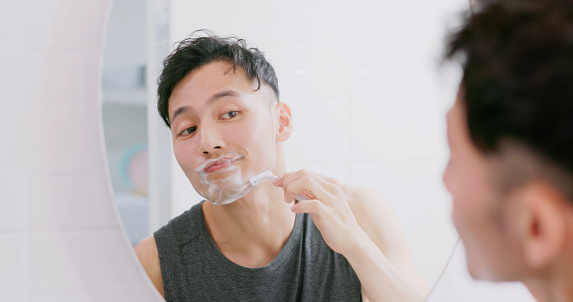 asian man standing in front of mirror is using razor and shaving face with foam