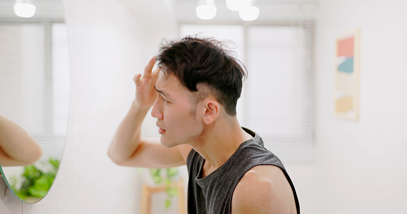 side view asian man standing in front of mirror is concerned about hair loss or alopecia