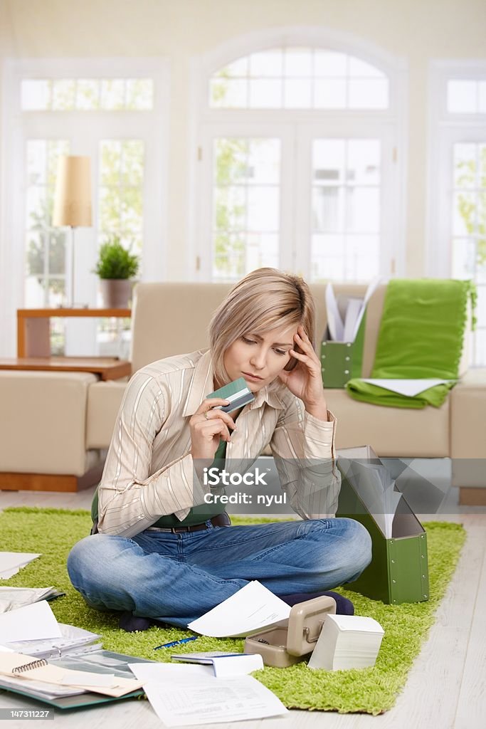 Troubled woman with credit card Troubled woman sitting on floor with crossed legs, looking at documents holding credit card in living room. 20-29 Years Stock Photo