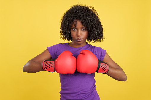 African woman clapping red boxing gloves while looking at camera in studio with yellow background