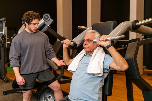 Portrait of senior man with fitness instructor pulling weight machine at the gym. The trainer teaches how to do the full form of the movement