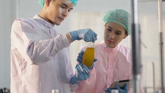 Asian male engineers or technicians and and female nutritionist or quality control officer wear sterile clothing to prevent contamination, working in bottled fruit juice factory, check production and product quality of juice filling machine in factory.